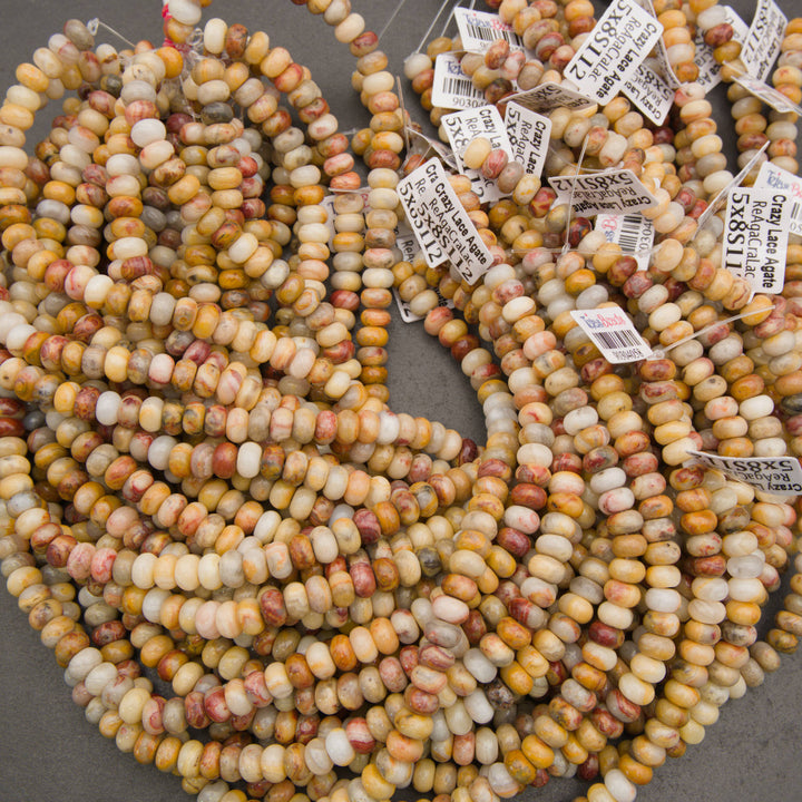Multiple strands of cylindrical marbled beads in warm yellow, orange, cream, and reddish-brown tones, with small product tags attached, photographed on a gray surface.