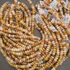 Multiple strands of cylindrical marbled beads in warm yellow, orange, cream, and reddish-brown tones, with small product tags attached, photographed on a gray surface.