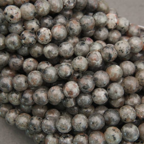 Multiple strands of polished round gemstone beads in earthy gray, brown, black, and white marbled patterns, with white inventory tags attached, photographed on a gray surface.