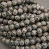 Multiple strands of polished round gemstone beads in earthy gray, brown, black, and white marbled patterns, with white inventory tags attached, photographed on a gray surface.