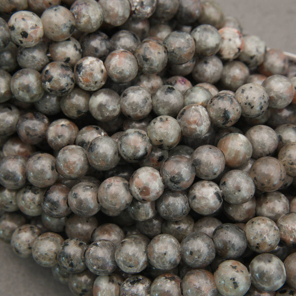 Multiple strands of polished round gemstone beads in earthy gray, brown, black, and white marbled patterns, with white inventory tags attached, photographed on a gray surface.