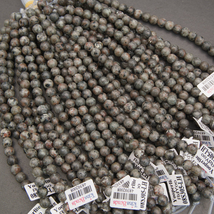 Multiple strands of polished round gemstone beads in earthy gray, brown, black, and white marbled patterns, with white inventory tags attached, photographed on a gray surface.