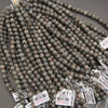 Multiple strands of polished round gemstone beads in earthy gray, brown, black, and white marbled patterns, with white inventory tags attached, photographed on a gray surface.