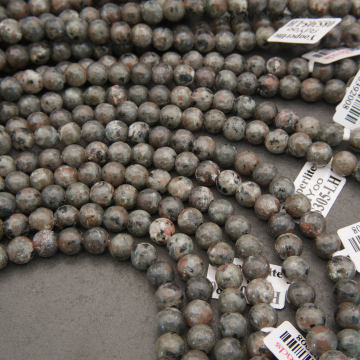 Multiple strands of polished round gemstone beads in earthy gray, brown, black, and white marbled patterns, with white inventory tags attached, photographed on a gray surface.