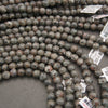 Multiple strands of polished round gemstone beads in earthy gray, brown, black, and white marbled patterns, with white inventory tags attached, photographed on a gray surface.