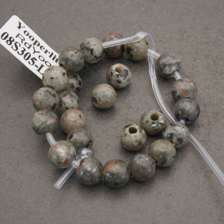 A strand of round natural stone beads with holes, displayed on a clear plastic tube. The beads have a mottled, earthy coloration of gray, brown, and beige with dark speckles, set against a neutral gray background.