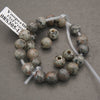A strand of round natural stone beads with holes, displayed on a clear plastic tube. The beads have a mottled, earthy coloration of gray, brown, and beige with dark speckles, set against a neutral gray background.