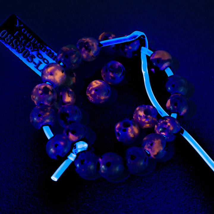 A set of UV-reactive beaded bracelets shown under blue light, revealing a purple and pink marbled bead design. One bracelet is laid flat, while another is wrapped around a white plastic loop to display its glow-in-the-dark properties.